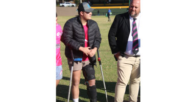 Young man stands on crutches with a leg brace, dressed casually, talking to a man in a suit on a sports field. Scene conveys support and teamwork.