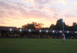 A rugby player prepares to kick a ball on a grassy field at dusk. The sky glows with a vibrant pink and orange sunset, and spectators sit in dimly lit stands.