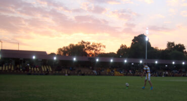 A rugby player prepares to kick a ball on a grassy field at dusk. The sky glows with a vibrant pink and orange sunset, and spectators sit in dimly lit stands.