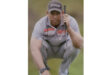 A golfer in a gray and orange polo and cap crouches intently on a green, holding a putter. The scene is focused and contemplative.