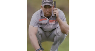 A golfer in a gray and orange polo and cap crouches intently on a green, holding a putter. The scene is focused and contemplative.