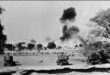 Black and white wartime scene with military vehicles in the foreground, smoke rising in the distance. Trees and open land convey a tense atmosphere.