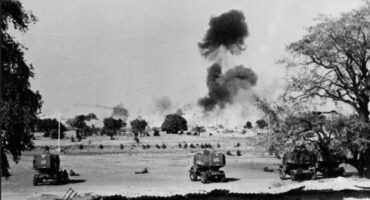 Black and white wartime scene with military vehicles in the foreground, smoke rising in the distance. Trees and open land convey a tense atmosphere.