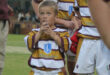 Young rugby player in a maroon and white striped jersey looks upwards, holding a water bottle. Other players in matching jerseys stand nearby on a grassy field.