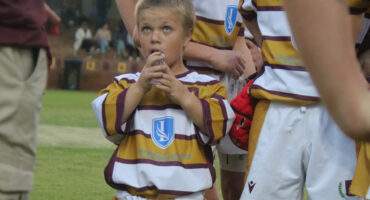Young rugby player in a maroon and white striped jersey looks upwards, holding a water bottle. Other players in matching jerseys stand nearby on a grassy field.