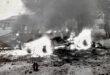 A black-and-white image depicts a burning vehicle in a desolate street. Flames and smoke rise dramatically, creating a chaotic and intense scene. Buildings and two figures are visible in the background.
