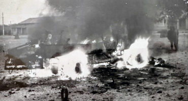 A black-and-white image depicts a burning vehicle in a desolate street. Flames and smoke rise dramatically, creating a chaotic and intense scene. Buildings and two figures are visible in the background.