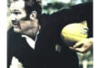 A rugby player in a black jersey holds a yellow ball tightly, focused and determined, with a blurred outdoor background suggesting action and movement.