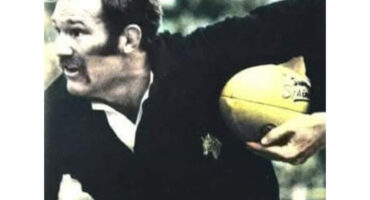 A rugby player in a black jersey holds a yellow ball tightly, focused and determined, with a blurred outdoor background suggesting action and movement.