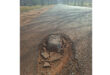 A deep pothole filled with rocks on a rural asphalt road, casting shadows. Surrounding trees and grass are visible, suggesting a neglected, worn path.