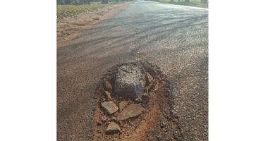 A deep pothole filled with rocks on a rural asphalt road, casting shadows. Surrounding trees and grass are visible, suggesting a neglected, worn path.