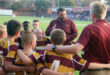 A youth rugby team in maroon and yellow jerseys huddles around a coach giving an animated pep talk on a green field, with a stadium in the background.