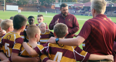 A youth rugby team in maroon and yellow jerseys huddles around a coach giving an animated pep talk on a green field, with a stadium in the background.