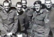 A group of seven smiling military men in uniform stand together outdoors. The man on the right wears a beret. The tone is camaraderie and friendship.