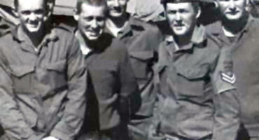 A group of seven smiling military men in uniform stand together outdoors. The man on the right wears a beret. The tone is camaraderie and friendship.