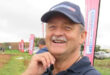 Smiling man in a navy cap outdoors at an event, with pink banners and parked cars in the background. Overcast sky suggests a cheerful, casual atmosphere.