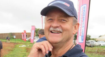 Smiling man in a navy cap outdoors at an event, with pink banners and parked cars in the background. Overcast sky suggests a cheerful, casual atmosphere.