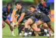 Rugby players in action on a grassy field; one player in black and gold prepares to pass the ball, while teammates and opponents focus intensely.