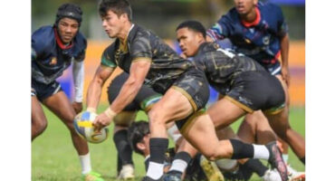 Rugby players in action on a grassy field; one player in black and gold prepares to pass the ball, while teammates and opponents focus intensely.