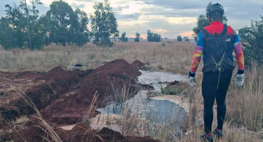 A person in colorful cycling gear and helmet stands in a grassy field at dusk, peering at a water-filled trench with trees in the background.