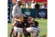 A rugby coach in a hat stands beside a huddled group of players in brown and white uniforms on a field. The scene is intense and supportive.