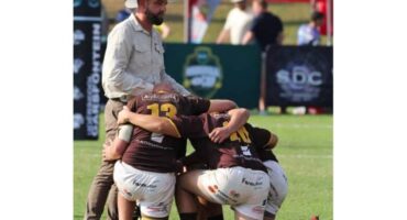 A rugby coach in a hat stands beside a huddled group of players in brown and white uniforms on a field. The scene is intense and supportive.