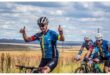 Three cyclists in matching blue jerseys ride through a grassy landscape. The lead cyclist smiles and gestures thumbs-up, conveying excitement and camaraderie.