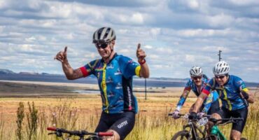 Three cyclists in matching blue jerseys ride through a grassy landscape. The lead cyclist smiles and gestures thumbs-up, conveying excitement and camaraderie.