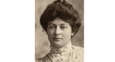 Portrait of a woman from the early 1900s with dark hair in an updo. She wears an embroidered, high-collared dress. The expression is calm and poised.