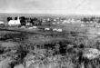 Black and white photo of a rural 1800s village. A prominent church is surrounded by fields and scattered houses, creating a serene, historical atmosphere.