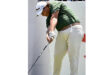 A man in a green shirt and white pants swings a golf club on a sunny golf course.