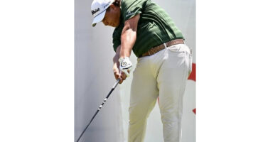A man in a green shirt and white pants swings a golf club on a sunny golf course.