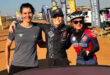 Three cyclists smiling with arms around each other at an outdoor event. They're wearing cycling gear, and banners are visible in the background.