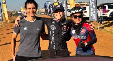 Three cyclists smiling with arms around each other at an outdoor event. They're wearing cycling gear, and banners are visible in the background.