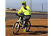 A cyclist wearing a neon green helmet and jacket rides on a dirt path near a roadway. The sunny outdoor setting conveys a sense of energy and enjoyment.
