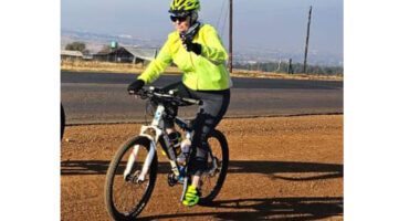 A cyclist wearing a neon green helmet and jacket rides on a dirt path near a roadway. The sunny outdoor setting conveys a sense of energy and enjoyment.
