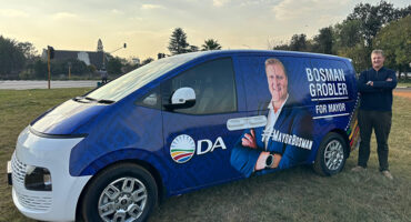 A man stands beside a blue campaign van on grass. The van displays a large image of another man with text "Bosman Grobler for Mayor" and "DA."