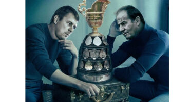 Two men are seated on a suitcase, holding a trophy, symbolizing their success and camaraderie.