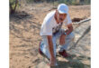 A man in a white cap and T-shirt kneels on a dusty archaeological site, examining a stone surface with a small tool. The setting is outdoors, under bright sunlight.