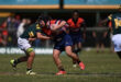 A rugby player in a blue and red uniform runs forward, holding the ball tightly, while another player in green and yellow attempts a tackle on a sunny field.