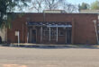 A small, brick building with a flat roof and two metal doors flanking a barred window. The setting is quiet, with trees and a clear sky above.