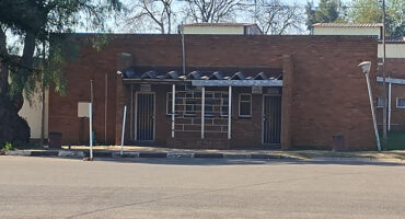 A small, brick building with a flat roof and two metal doors flanking a barred window. The setting is quiet, with trees and a clear sky above.