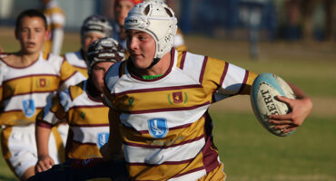 A rugby player in a striped jersey and helmet runs with determination, holding a rugby ball. Teammates follow closely on a sunlit field.