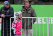 A young rugby player in a pink jersey and protective headgear stands on the field, focused. Two spectators are in the background near a green sign.