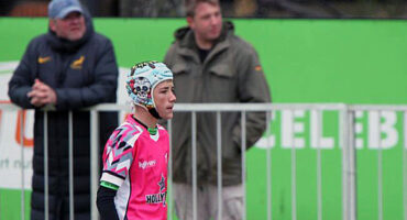 A young rugby player in a pink jersey and protective headgear stands on the field, focused. Two spectators are in the background near a green sign.