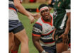 Rugby players in action; one, wearing a red and white jersey with a headband, looks intensely focused. They’re surrounded by teammates on a grassy field.
