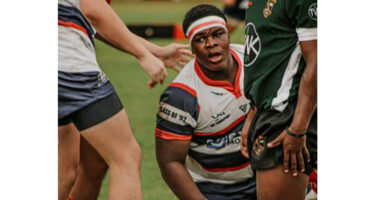 Rugby players in action; one, wearing a red and white jersey with a headband, looks intensely focused. They’re surrounded by teammates on a grassy field.