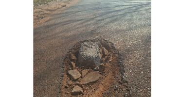 A large pothole filled with rocks on an asphalt road, casting shadows in the sunlight. The road appears worn and extends into the distance.