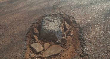 A large rock obstructs the center of a road, surrounded by asphalt and grass on either side.