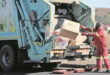 A man in a red shirt is loading trash into a garbage truck on a city street.